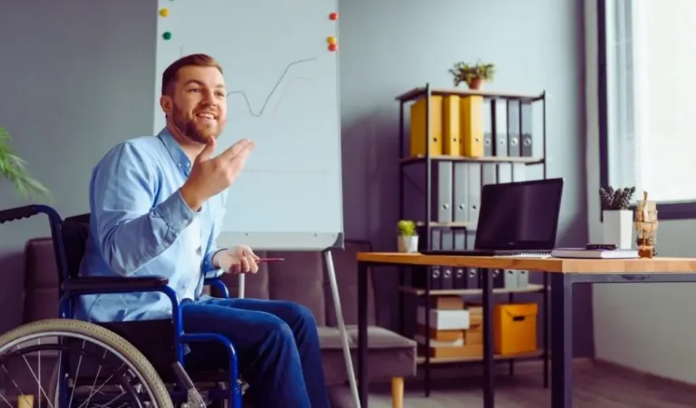 Na zdjęciu widoczny jest uśmiechnięty mężczyzna na wózku inwalidzkim, który prowadzi prezentację w nowoczesnym biurze. Za nim stoi tablica z wykresem, a obok znajduje się biurko z laptopem, książkami i roślinami. W tle widać regał z kolorowymi segregatorami oraz duże okno wpuszczające naturalne światło. Atmosfera miejsca jest ciepła, profesjonalna i sprzyjająca zaangażowaniu. Zdjęcie oddaje poczucie pewności siebie, aktywności zawodowej i integracji. To przykład, jak wygląda dostępne środowisko pracy dla niepełnosprawnych – przestrzeń funkcjonalna, estetyczna i wspierająca rozwój każdej osoby.